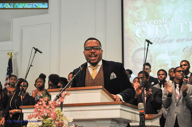 A man in a suit and tie speaking at a pulpit with people in the background.