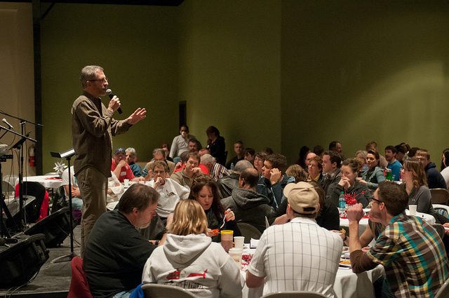 A man on a stage with a microphone speaking to a group of people in an informal setting.