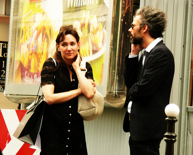 Two people standing together, the woman is looking away with an awkward expression, and the man is looking away and on his phone.