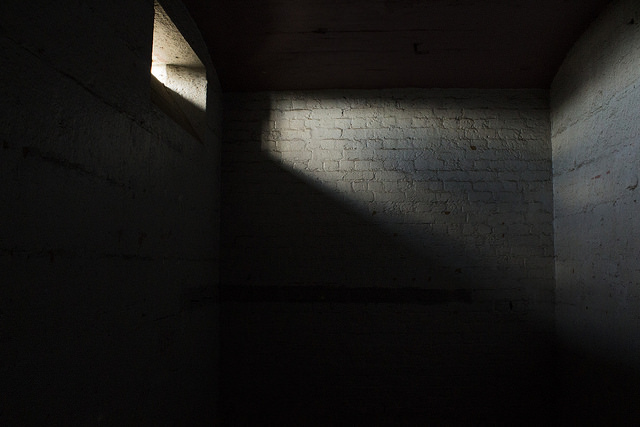 A small dark brick cell with limited light coming through a small window near the ceiling.