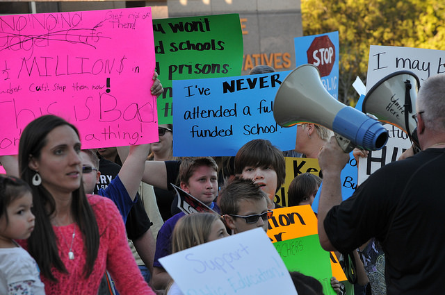 Adults and children at a rally with signs, one reading "I've never attended a fully funded school."
