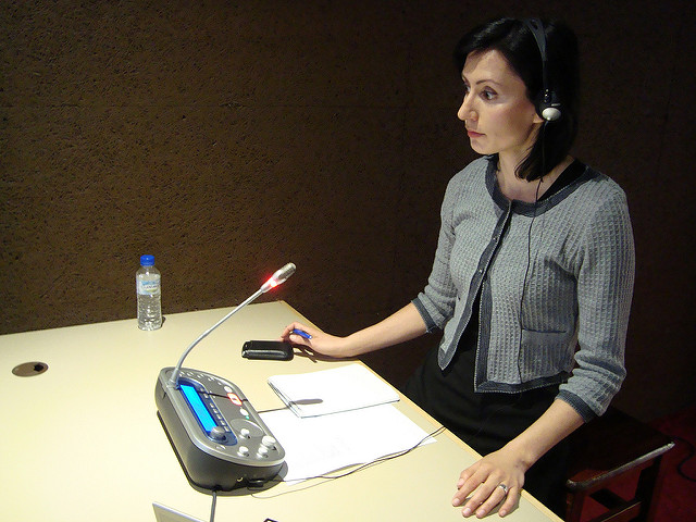 A female interpreter at a desk wearing headphones with a microphone in front of her.