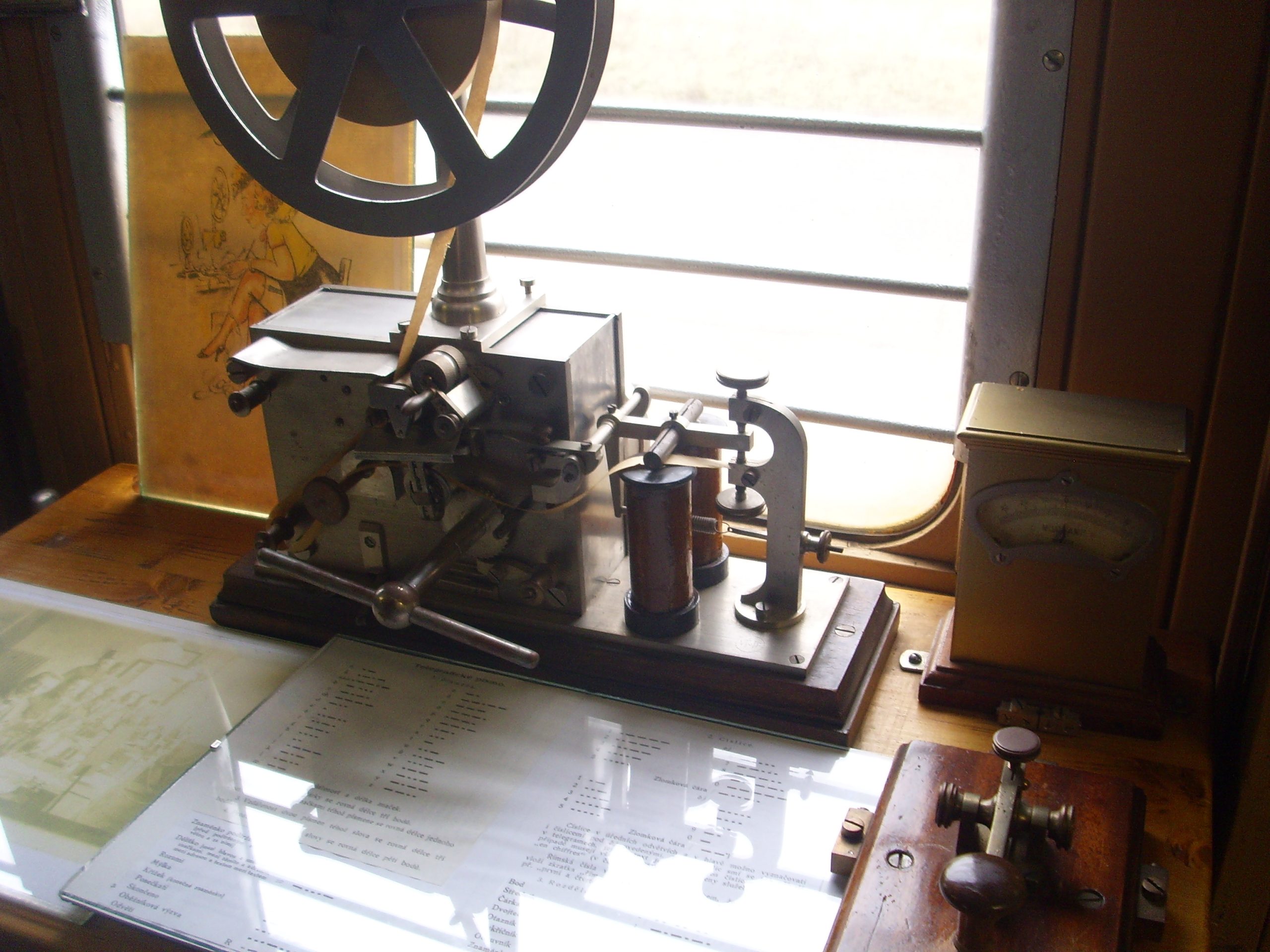A vintage electrical telegraph machine with levers, a wheel, and a gauge on a wooden desk.