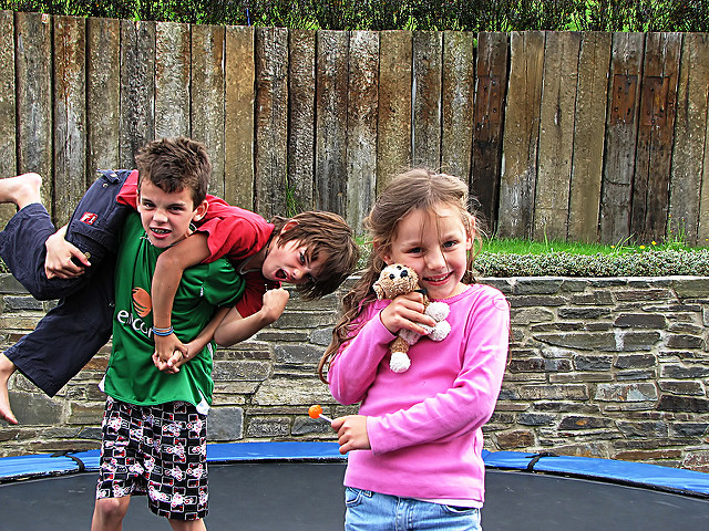 One young boy holding another boy on his back while a young girl wearing a pink shirt poses with a teddy bear and sucker.