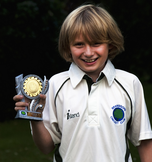 A smiling young boy wearing a shirt with the words "Bransgore Cricket Club" and holding an award.