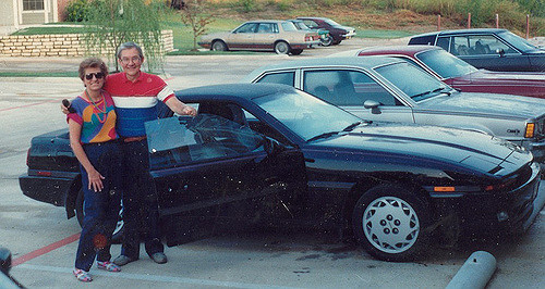 An older man and woman standing in front of a sports car.