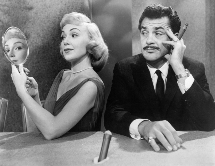 Two people sitting side by side with the woman's image being reflected in a hand held mirror, and the man looking sideways at her.