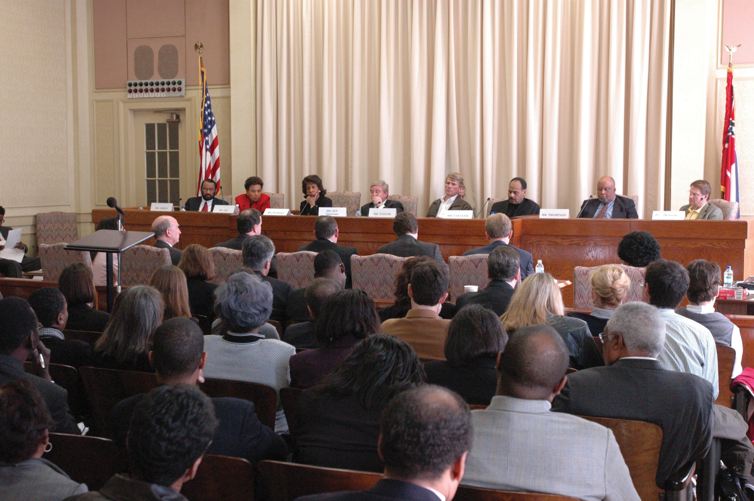 Congressional hearing held in a large room with eight people sitting behind a long desk with 2 flags behind the desk, and people in the audience.