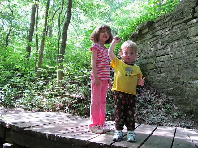 A male toddler holding a rock agressively overhead ready to throw it. A young girl standing behind him with a sneering look on her face.