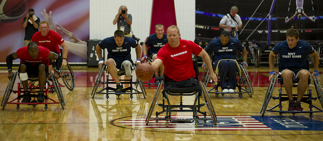 American veterans in red and blue shirts playing basketball in wheelchairs.
