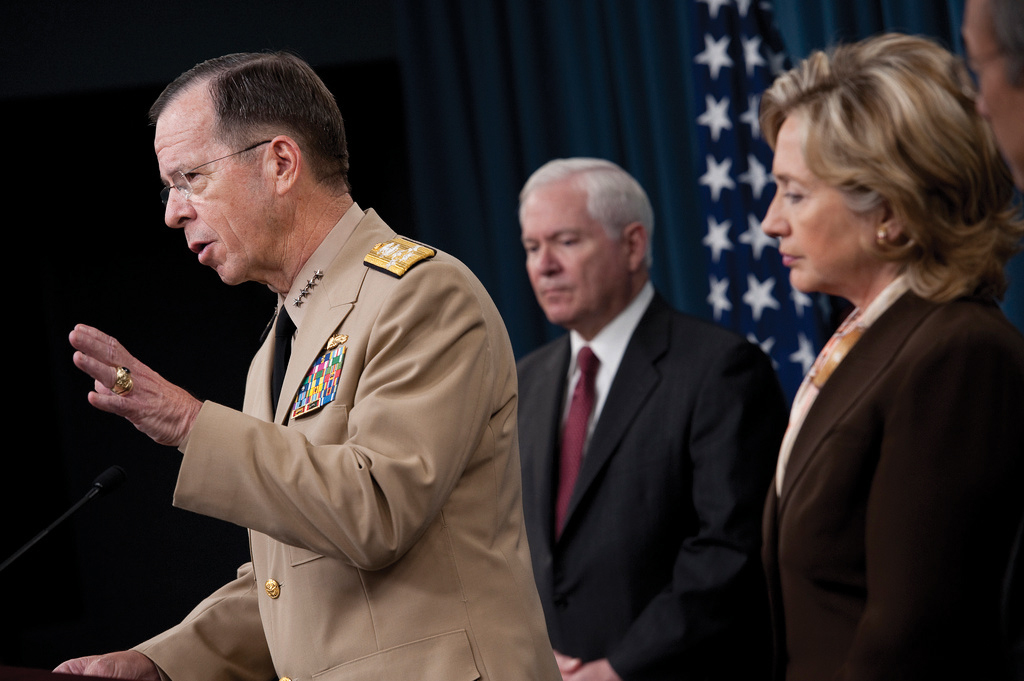 A military leader speaking behind a podium with three people behind him including Hillary Clinton. All of them are standing up straigh