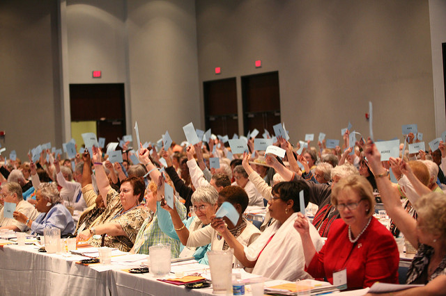 Women seated in a large ballroom looking engaged and holding up blue strips of paper.